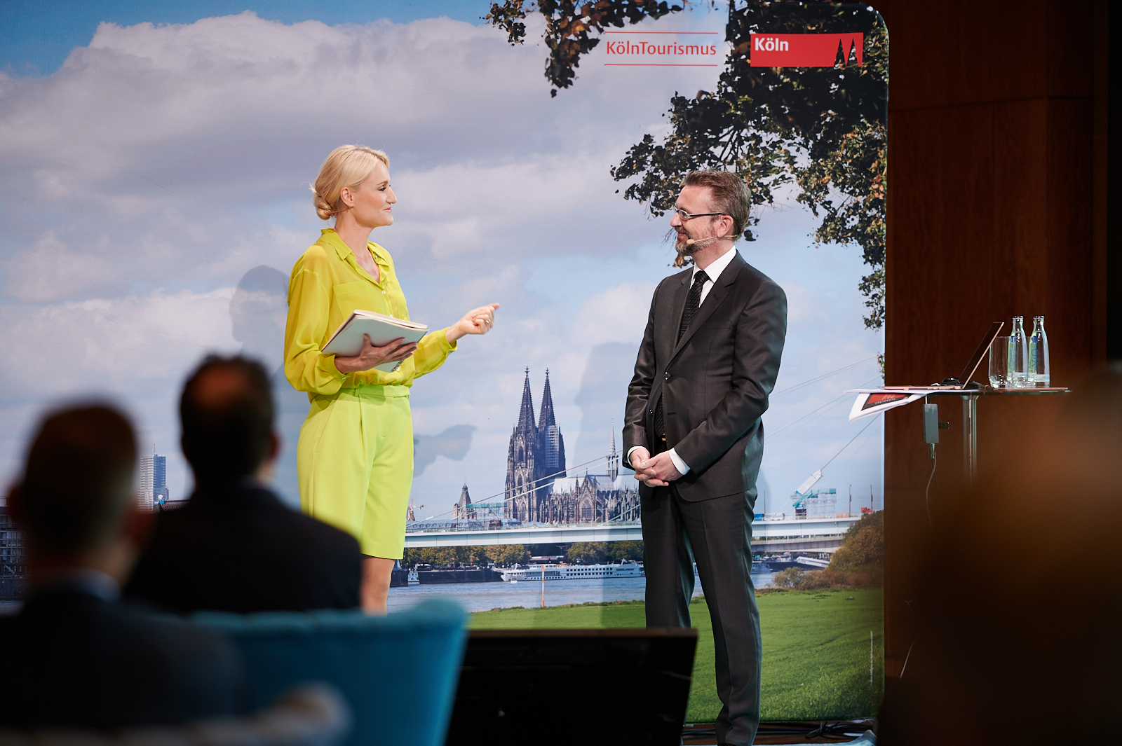 Zwei Personen unterhalten sich auf der Bühne, im Hintergrund eine Stellwand mit Köln Panorama