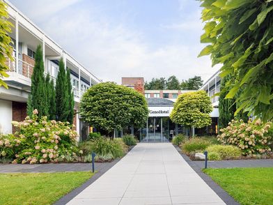 Hoteleingang des GenoHotel Forsbach mit grüner Rasenfläche und Bäumen