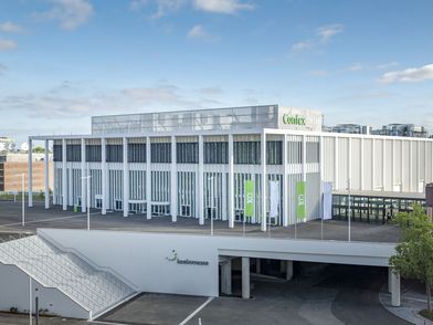Neues Kongresszentrum Confex von außen: Ein weißes, großes, modernes Gebäude mit einer Glasfront und hohen Säulen davor, außerdem Treppen, die zum erhöhten Gebäude führen.