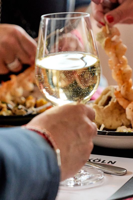 Eine Hand mit einem Glas Weißwein und zwei Teller mit Essen im Hintergrund