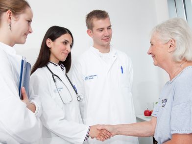 Ein Ärzteteam von zwei Ärztinnen und einem Arzt begrüßt eine ältere Dame als Patientin mit Handschütteln