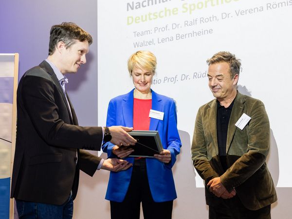 Ein Mann übergibt zwei anderen Männern der Deutschen Sporthochschule den Humboldt Preis