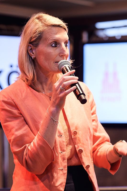 Eine Frau in einem orangenen Blazer hält eine Rede mit einem Mikrofon