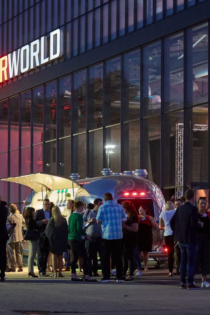 Motorworld Köln von außen. Vor der großen Halle stehen Menschen, die sich unterhalten.
