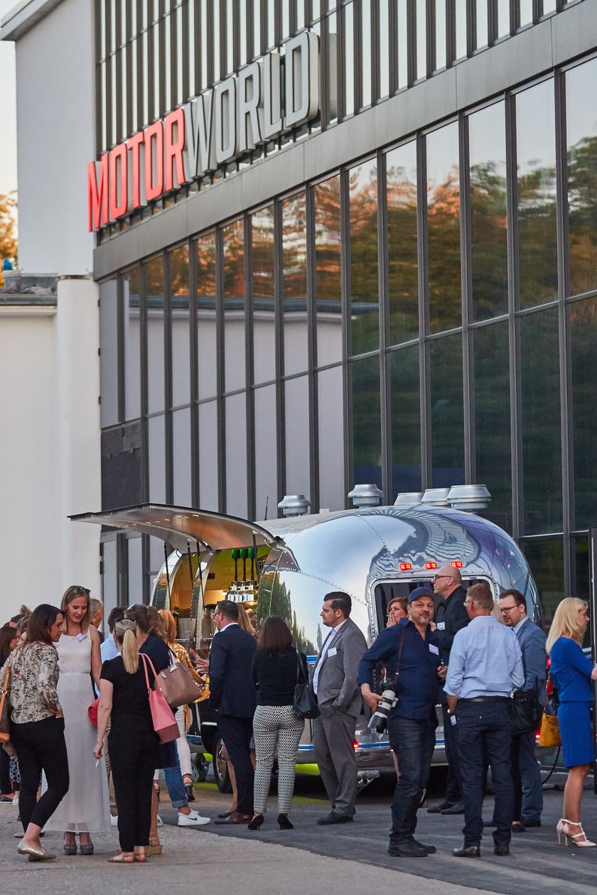 Motorworld von außen. Vor der Halle stehen Personen, die sich unterhalten.