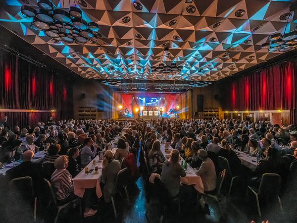 Veranstaltung in der Stadthalle Köln: Menschen sitzen an Tischreihen und schauen auf das Bühnenprogramm