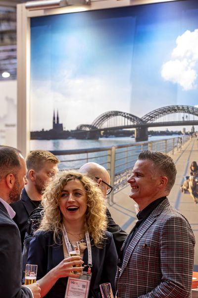 Personen trinken Kölsch vor dem Messestand des CCB und unterhalten sich