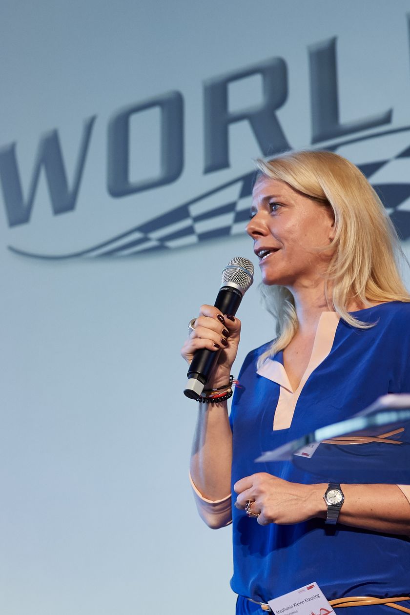 Frau hält eine Rede vor einer Leinwand mit der Aufschrift "Motorworld"