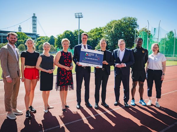 9 Menschen stehen in einer Reihe auf dem Sportplatz der Deutschen Sporthochschule Köln und halten eine Finanzierungszusage in der Hand