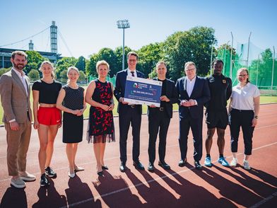 9 Menschen stehen in einer Reihe auf dem Sportplatz der Deutschen Sporthochschule Köln und halten eine Finanzierungszusage in der Hand